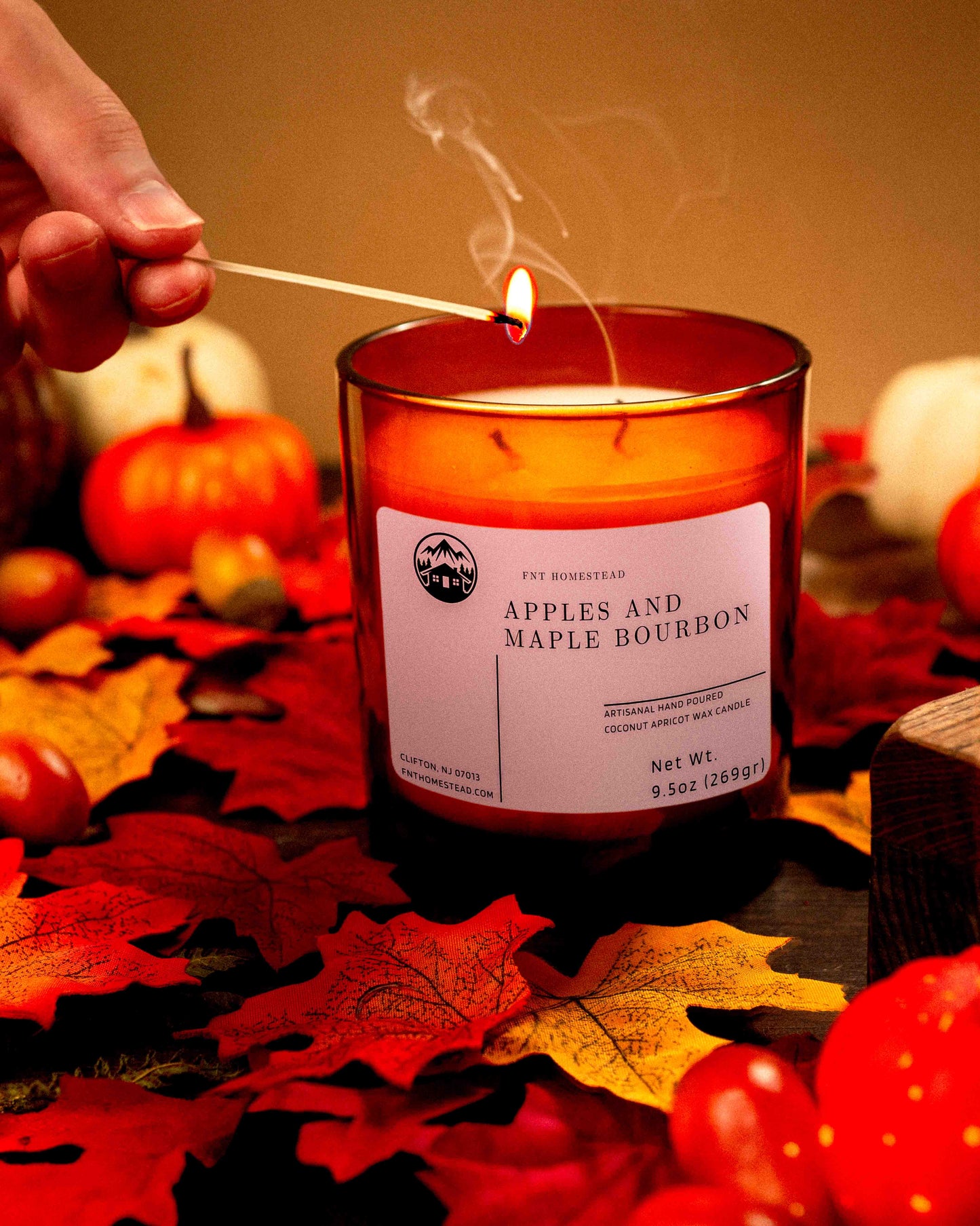 A person lights an amber jar candle labeled 'Apples and Maple Bourbon', surrounded by fall leaves and pumpkins."