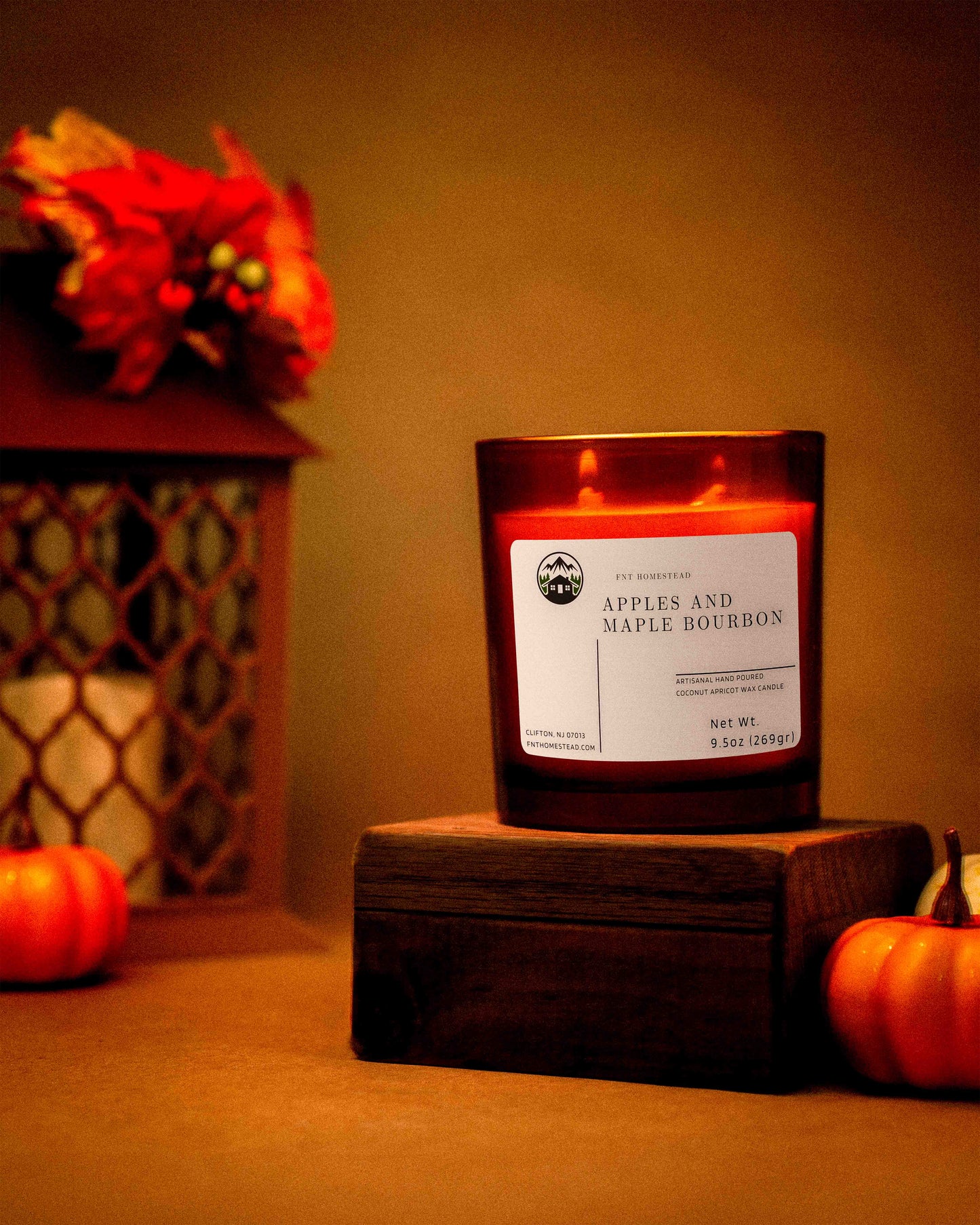 An amber jar candle labeled 'Apples and Maple Bourbon' sits on a wooden box, with two small pumpkins and a decorative lantern in the background. The warm lighting creates a cozy autumnal scene.