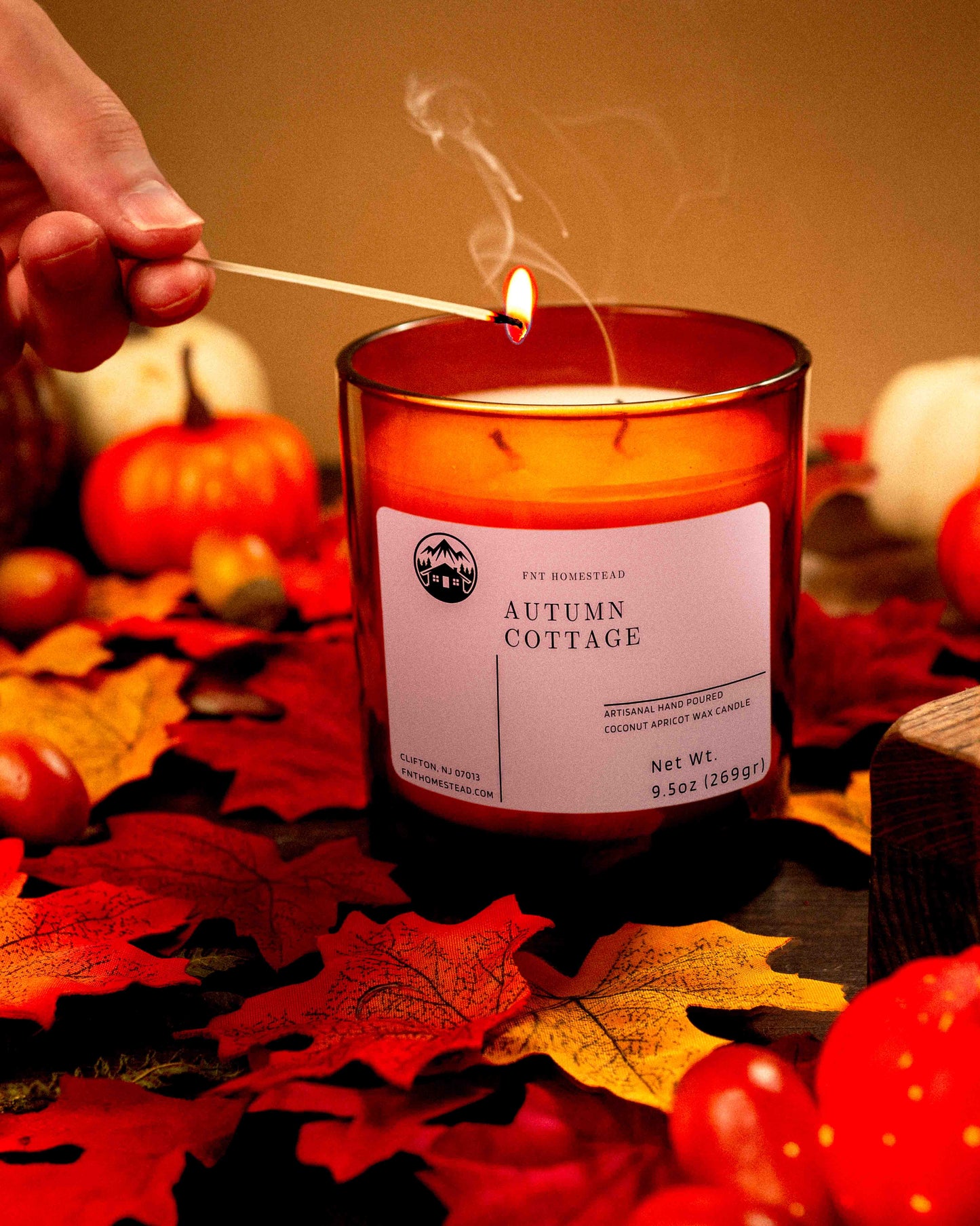 A person lights an amber jar candle labeled 'Autumn Cottage', surrounded by fall leaves and pumpkins.