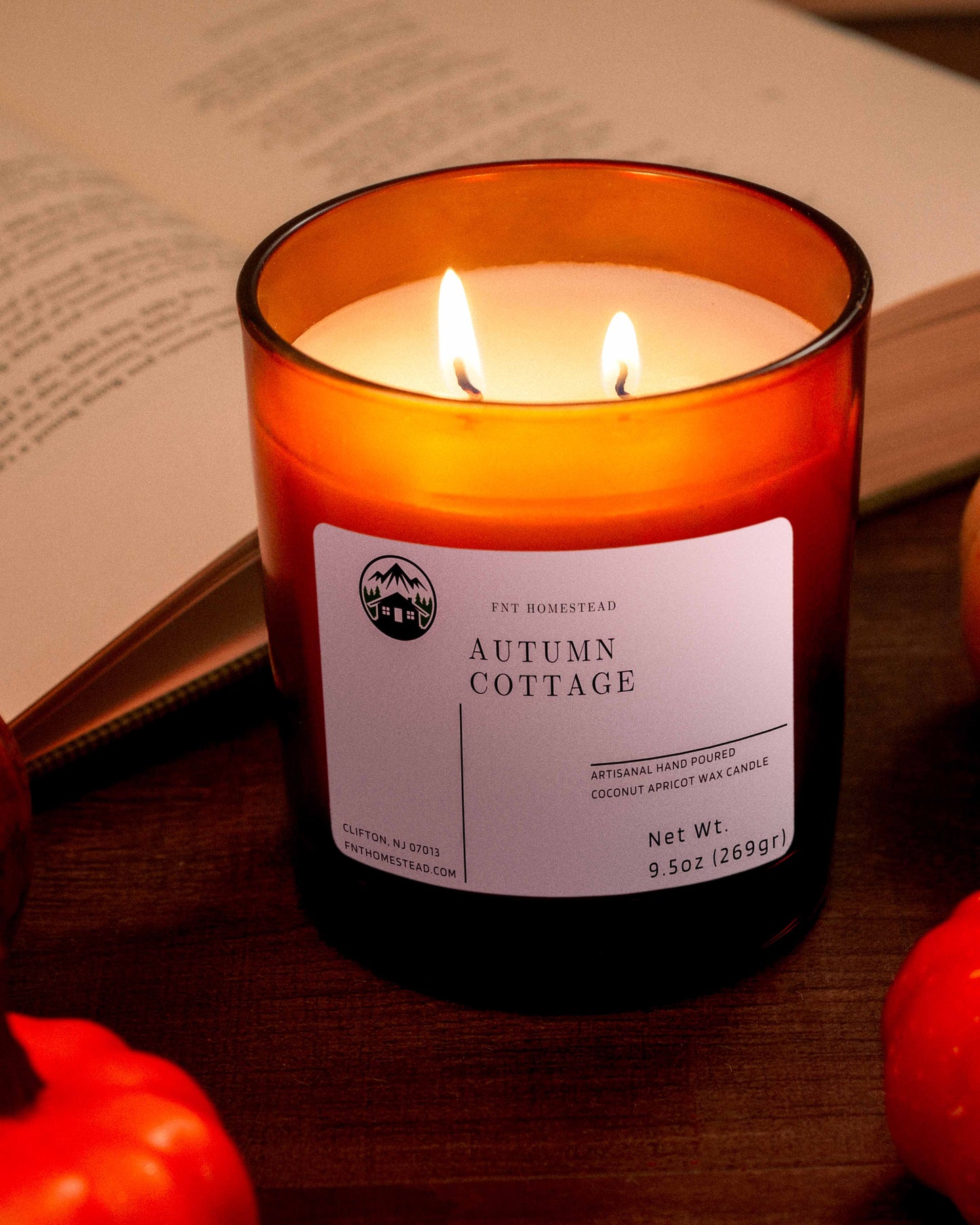 An amber jar candle with two lit wicks sits next to an open book. The candle is labeled 'Apples and Maple Bourbon' and has a fall-inspired scent. Three small pumpkins are placed near the candle.