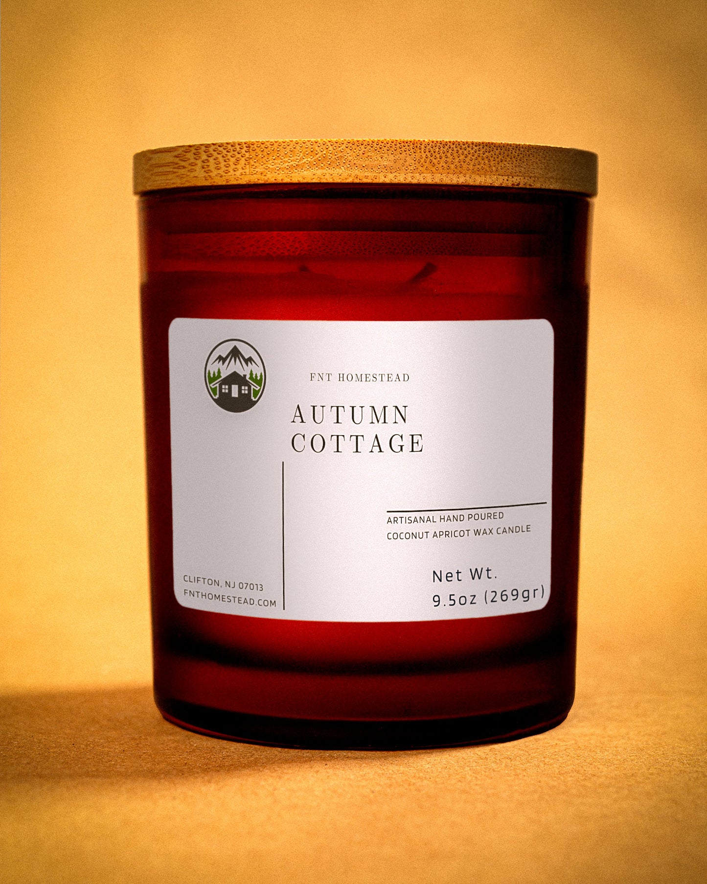 An amber jar candle with a wooden lid, labeled 'Autumn Cottage'. The label indicates it's an artisanal hand-poured candle made with coconut apricot wax.