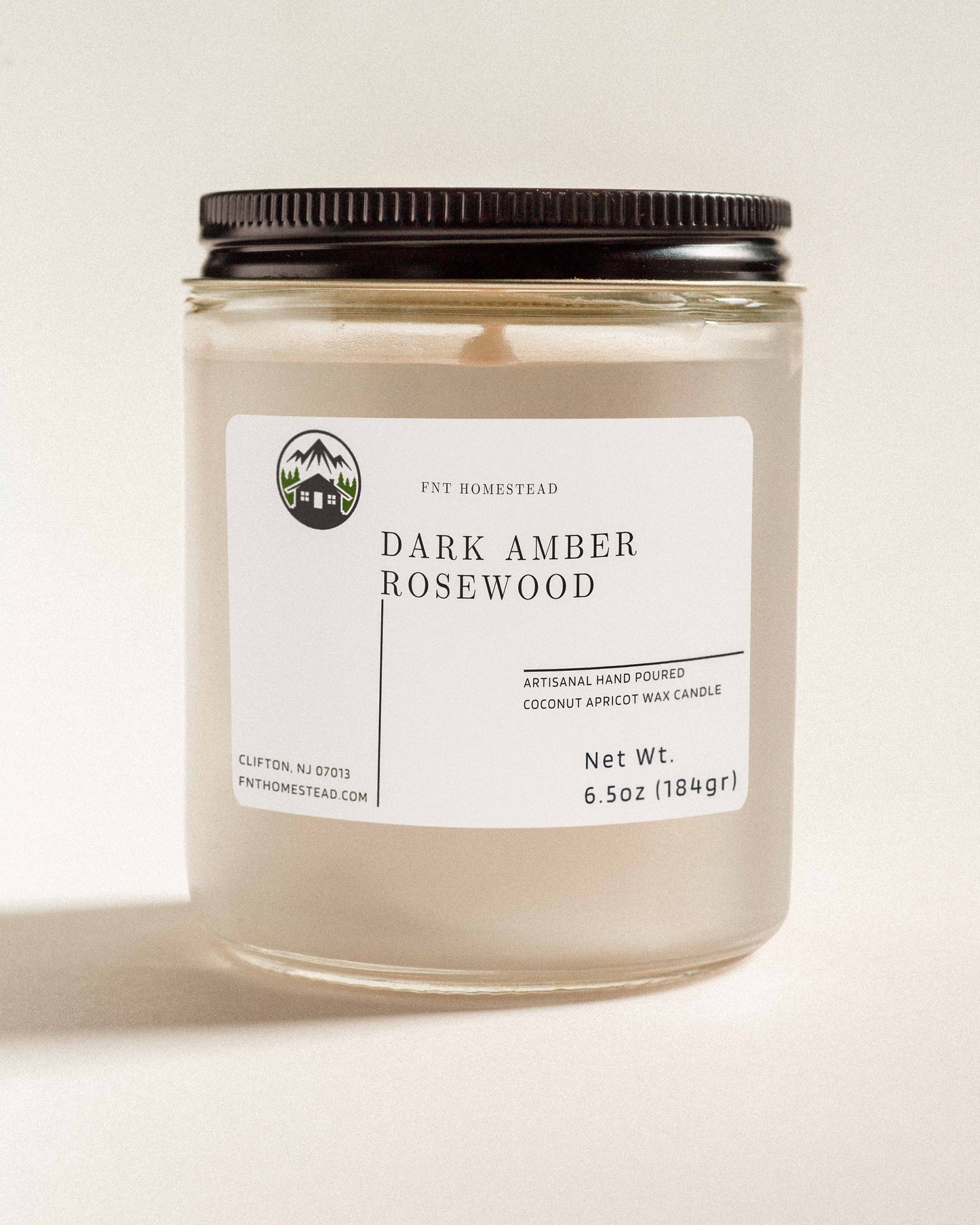 'Dark Amber Rosewood' scented candle in a clear glass jar with a black lid. This handcrafted candle features a smooth, white wax surface and is made with coconut apricot wax.