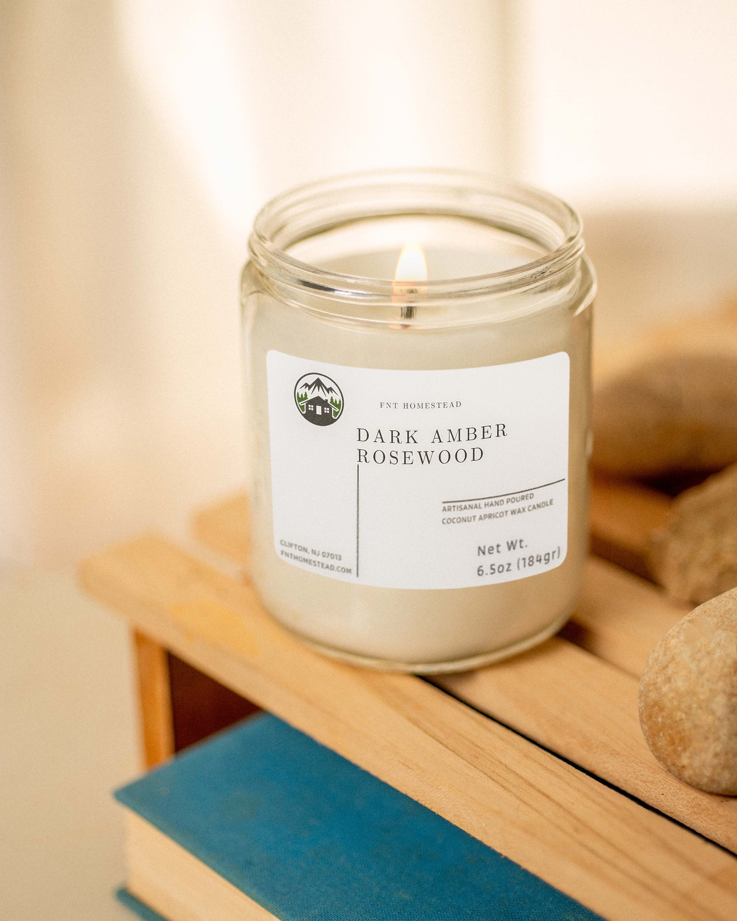 The 'Dark Amber Rosewood' candle creates a relaxing atmosphere with its soft glow in this clear glass jar, placed on a wooden surface with a book and stones.