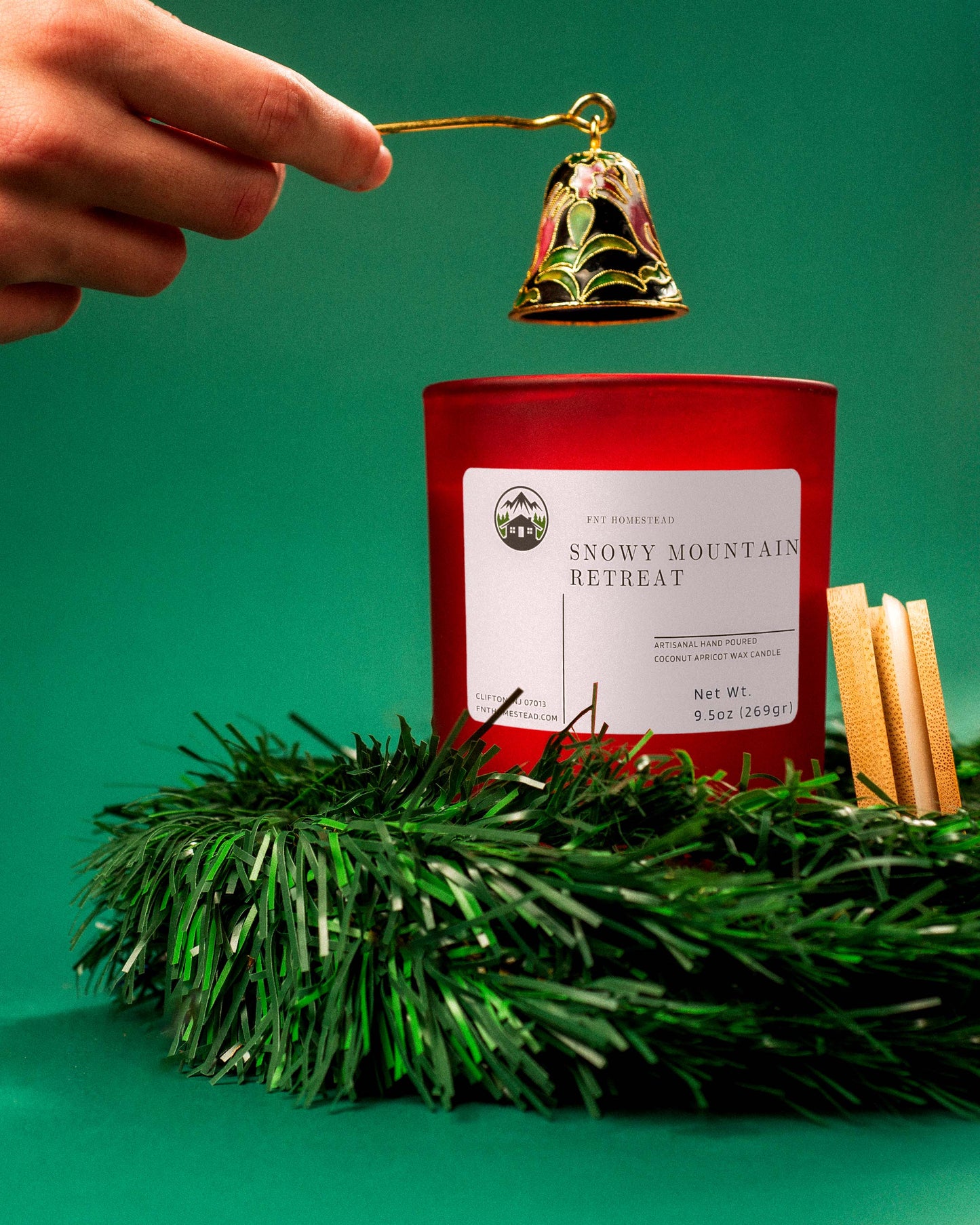 A hand holds a decorative candle snuffer over a red 'Snowy Mountain Retreat' candle. The candle sits in a wreath of evergreen branches, with the wooden lid sitting beside it.