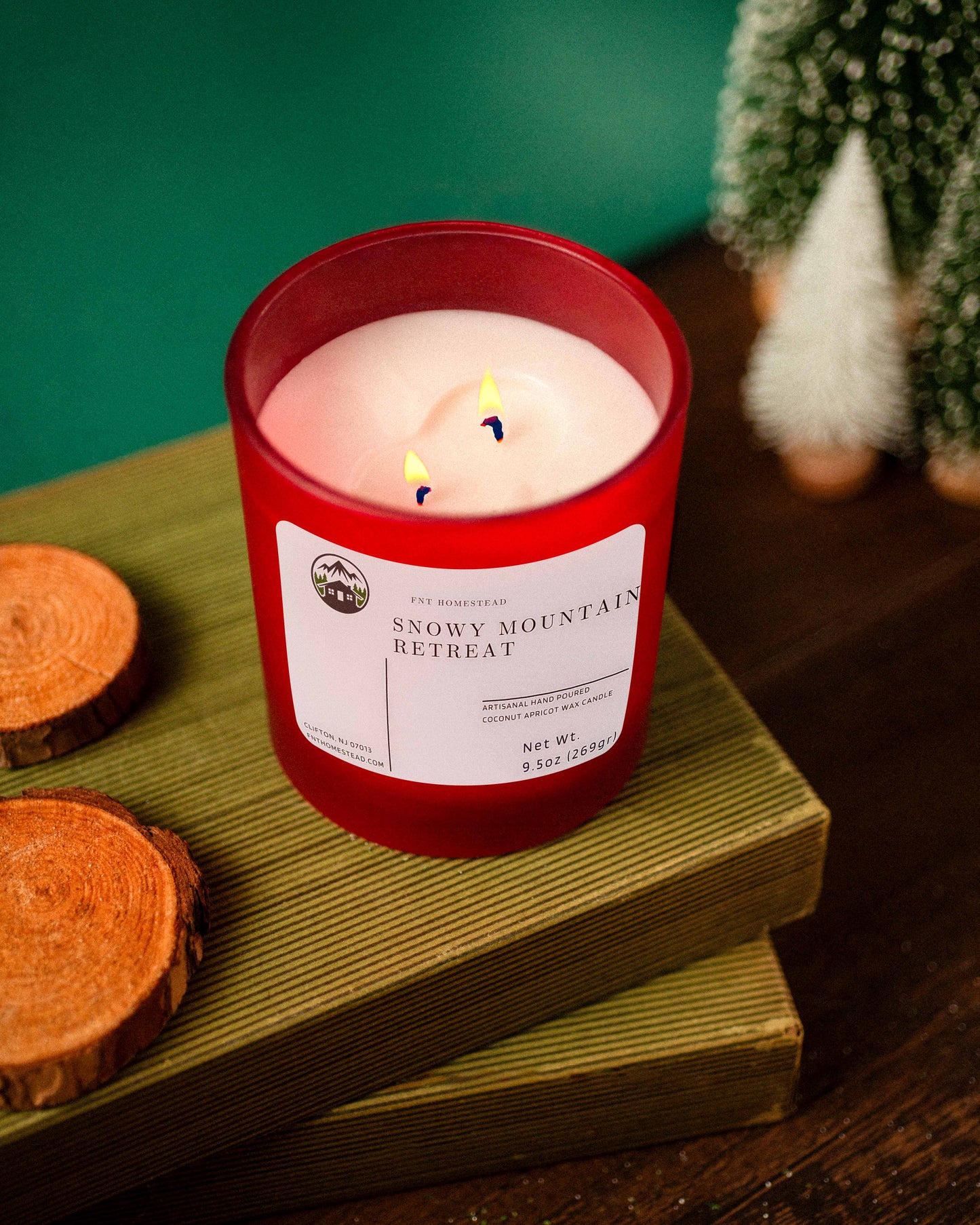 A red jar candle with two lit wicks sits on a stack of brown boards. The candle is labeled 'Snowy Mountain Retreat' and has a woodsy scent. Two wood slices are placed next to the boards, and a small decorative pine tree is in the background.