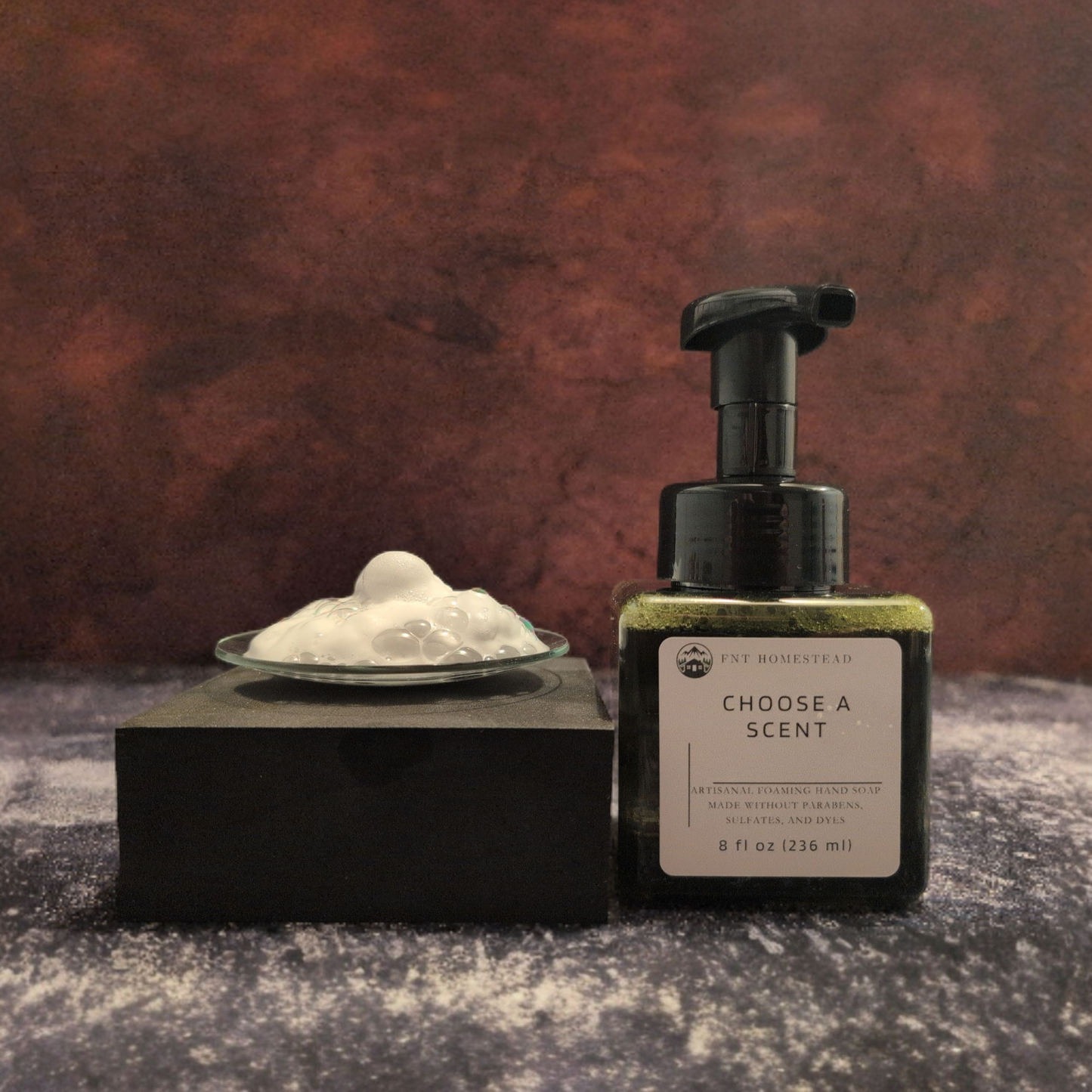 A bottle of FNT Homestead foaming hand soap is displayed next to a sample of its white foam. The dark green, square bottle with golden-hued soap is on the right. To the left, a swirl of white foam sits in a small, clear dish resting on a black block. Both items are set against a dark, textured, and moody background.