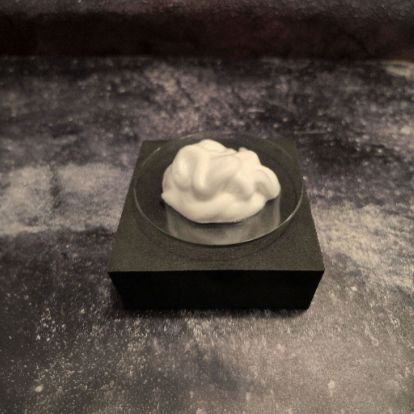 A swirl of white foam sits in a small, clear, round dish. The dish is centered on a solid black square block, which is placed on a dark, mottled gray and black surface.