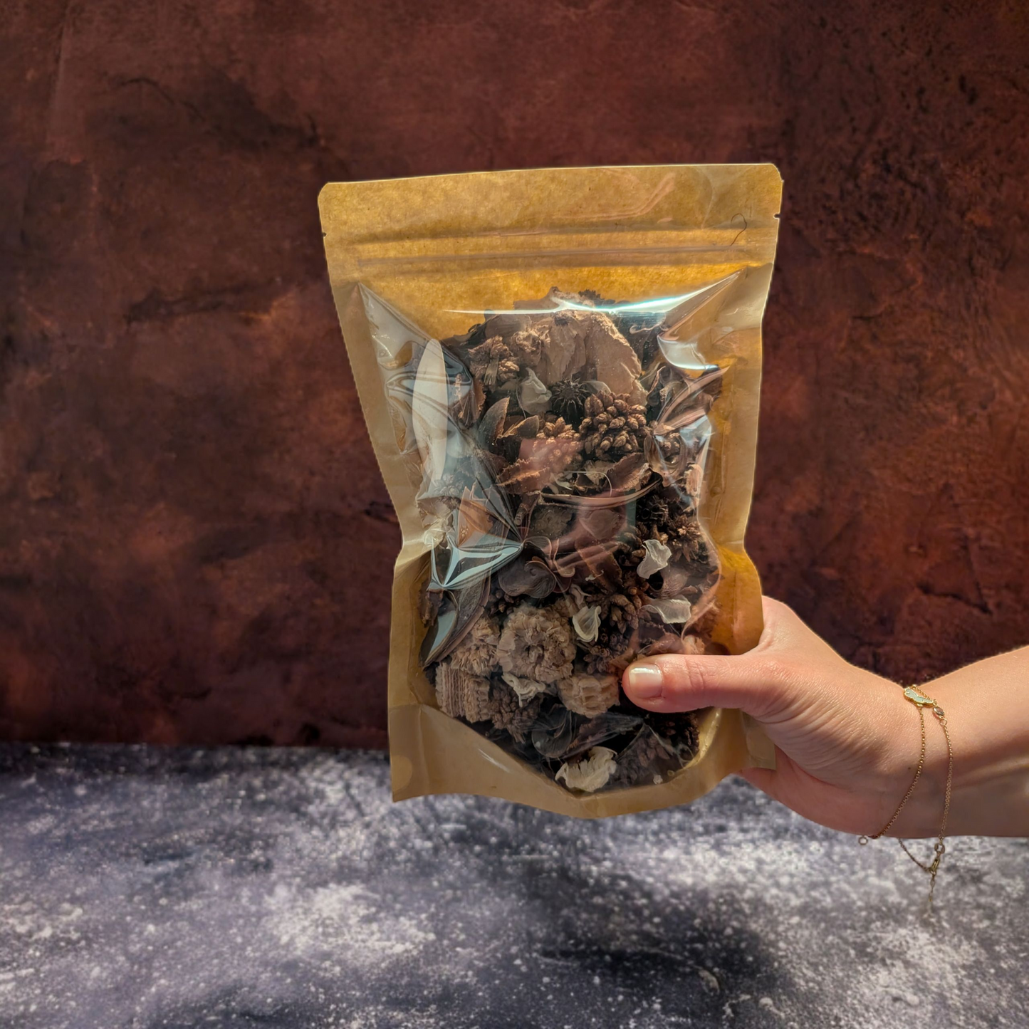 A person's hand holds a stand-up kraft paper pouch with a clear window, which is filled with brown and beige potpourri. The background is a dark, textured surface in shades of brown and gray.
