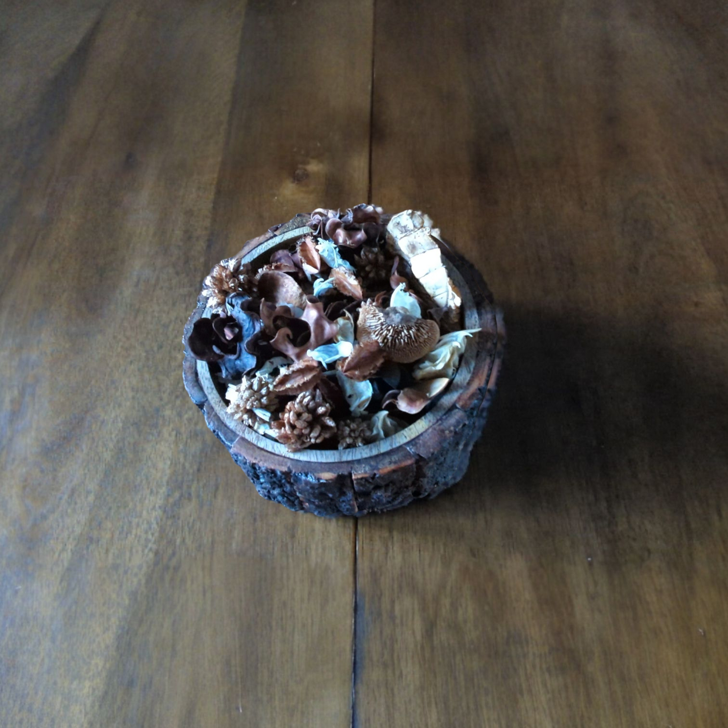 An overhead shot of a rustic bowl made from a tree log, filled with potpourri, sitting on a wooden table.