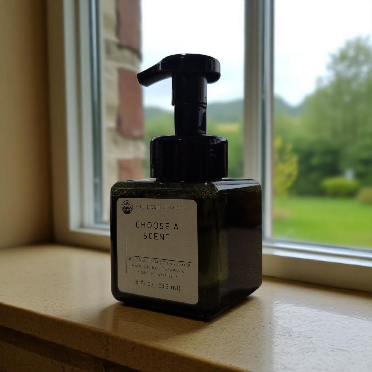 A dark green, square bottle of FNT Homestead foaming hand soap sits on a windowsill. The white label on the bottle says "Choose a Scent." Through the window behind the bottle is a soft-focus view of a green landscape.