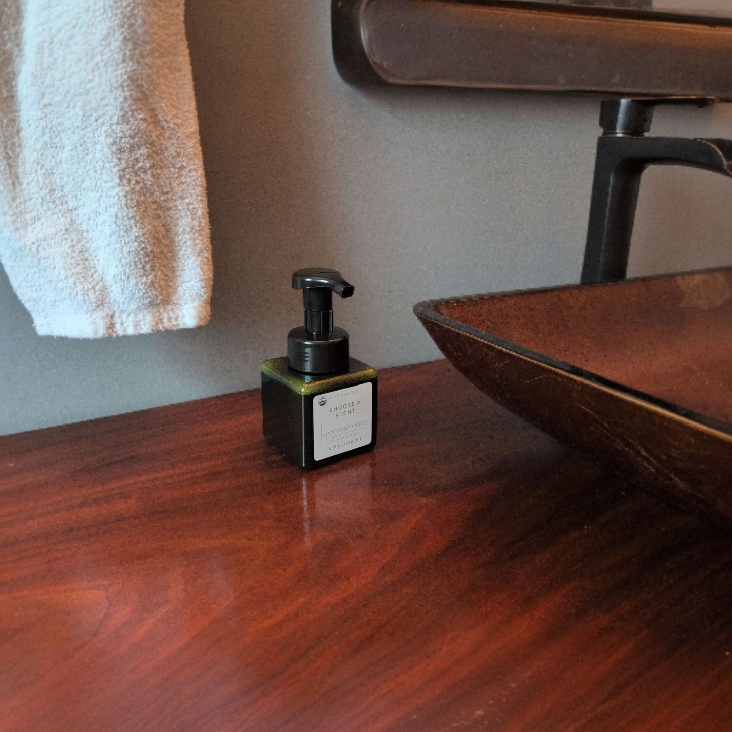 A dark green, square bottle of "Choose a Scent" hand soap sits on a dark wood bathroom counter. To the right is the edge of a textured, dark amber glass vessel sink, and to the left, a white hand towel hangs against the gray wall.