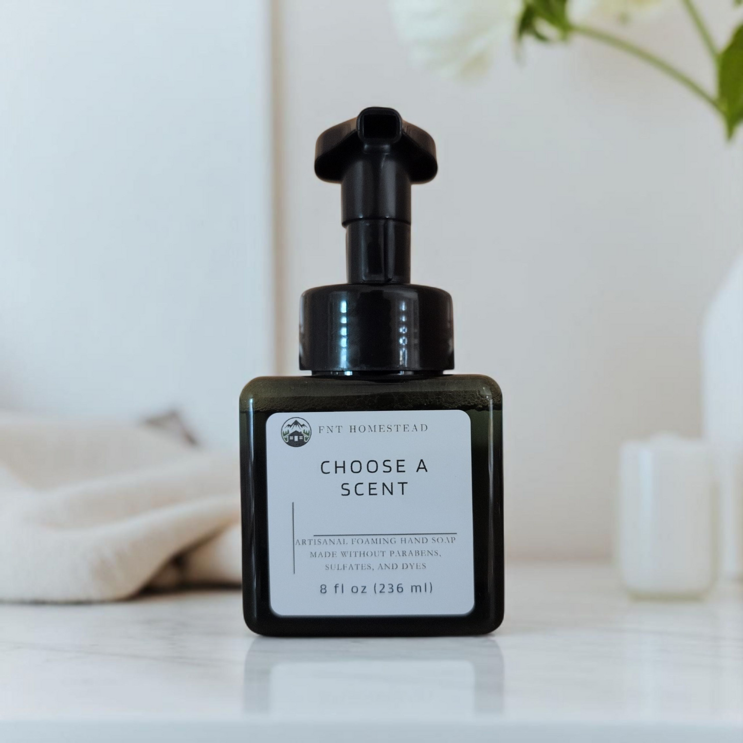 An angled, view of a dark green, square bottle of FNT Homestead foaming hand soap on a white surface. The label reads, "Choose a Scent." The bottle is flanked by a folded tan cloth on the left and a white vase with white flowers on the right, both of which are softly out of focus.