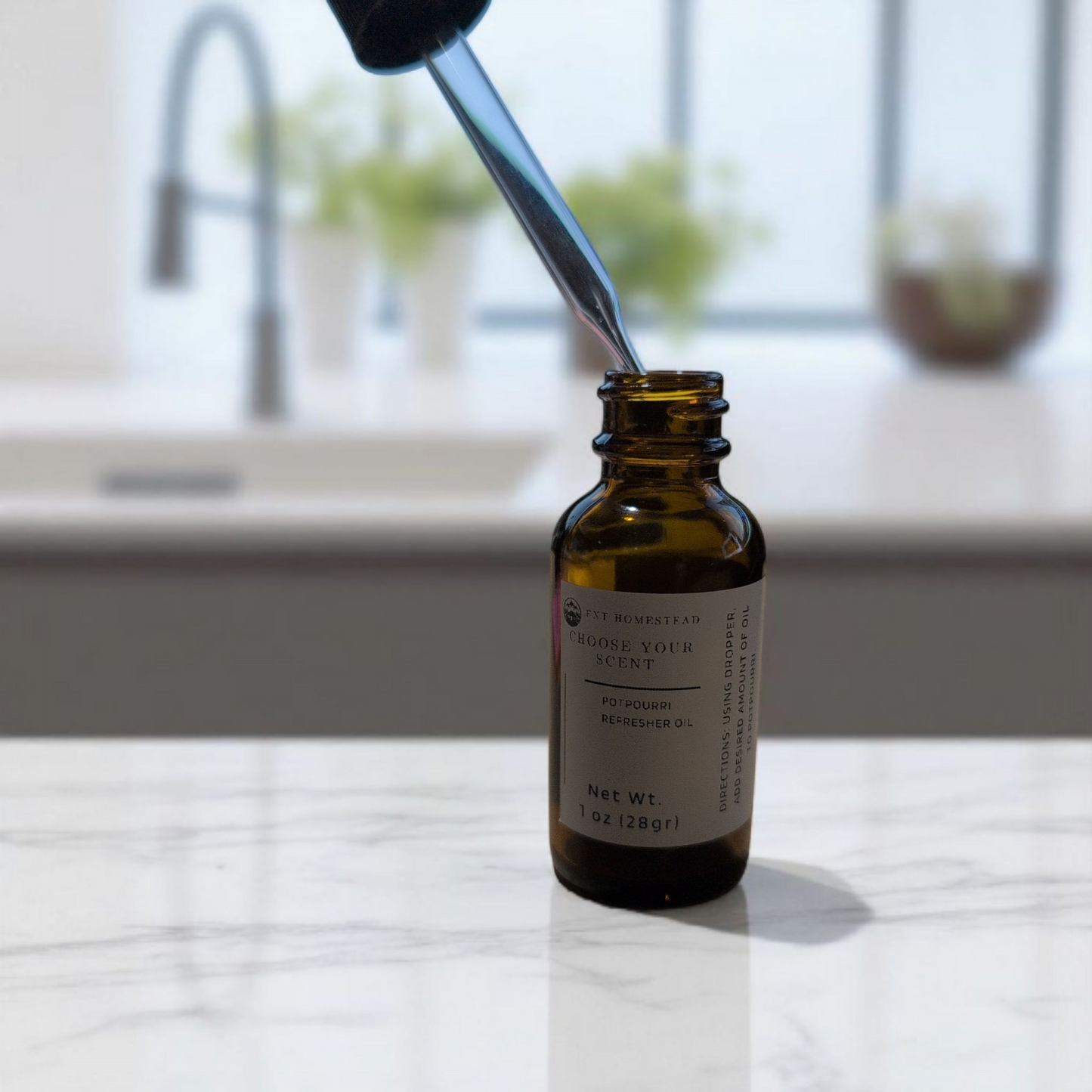 This is a bright, lifestyle photograph showing the product in a home setting.
An open, 1 oz. amber glass bottle of "Potpourri Refresher Oil" sits in the foreground on a white and grey marble countertop, which shows a soft reflection of the bottle.
The glass pipette of the dropper is angled down and is being inserted into the neck of the bottle. The background is a softly blurred, modern kitchen, with a gooseneck faucet, a window, and small, green potted plants visible behind the counter.