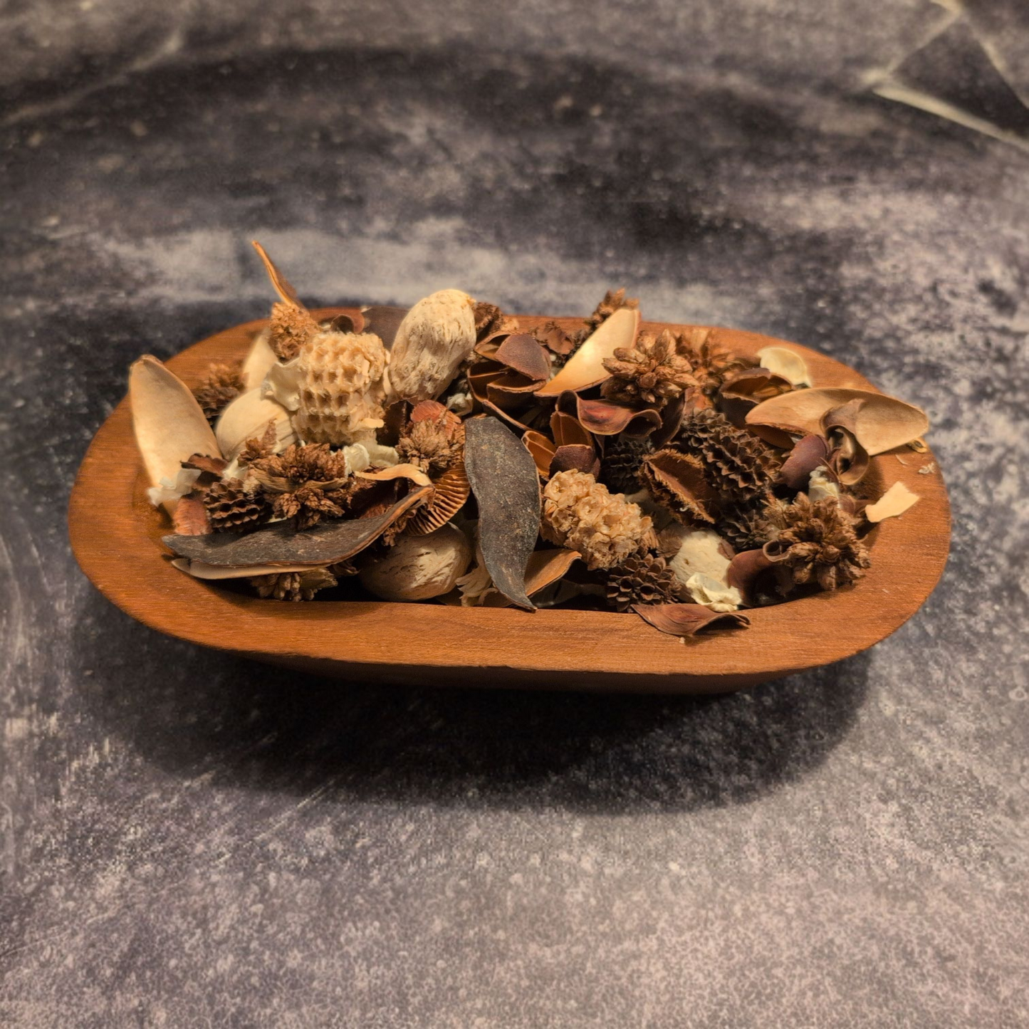 A shallow, oval-shaped wooden bowl filled with brown and cream-colored potpourri, sitting on a dark, textured gray surface.