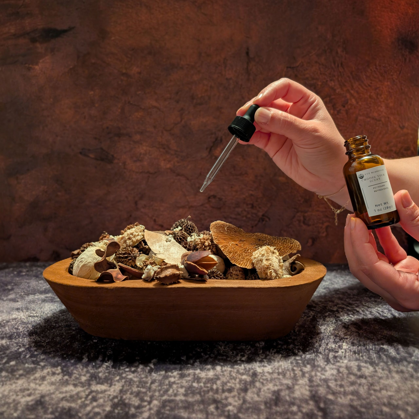 This is a warm, atmospheric photograph demonstrating a product in use.
The image shows a person's hands adding scented oil to a bowl of potpourri. One hand holds a 1 oz. amber glass bottle of "Potpourri Refresher Oil," while the other holds the dropper over a low, terracotta-colored bowl filled with dried botanicals.
A single drop of oil is captured in mid-air, falling from the glass pipette into the potpourri below. The scene is set on a dark, textured surface against a deep reddish-brown background.
