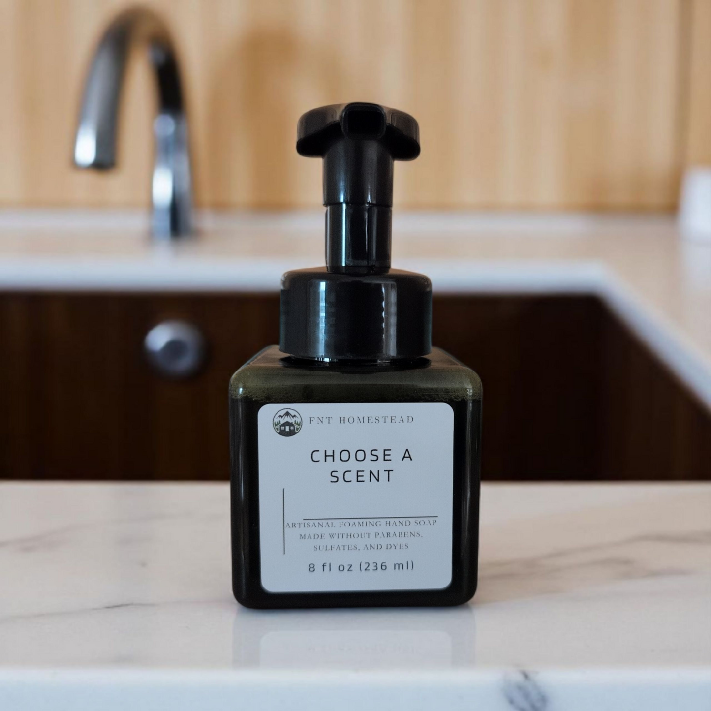 A dark green, square bottle of FNT Homestead foaming hand soap sits on a white and grey marble countertop. In the blurred background is a kitchen sink and a silver faucet. The label on the bottle reads, "Choose a Scent."