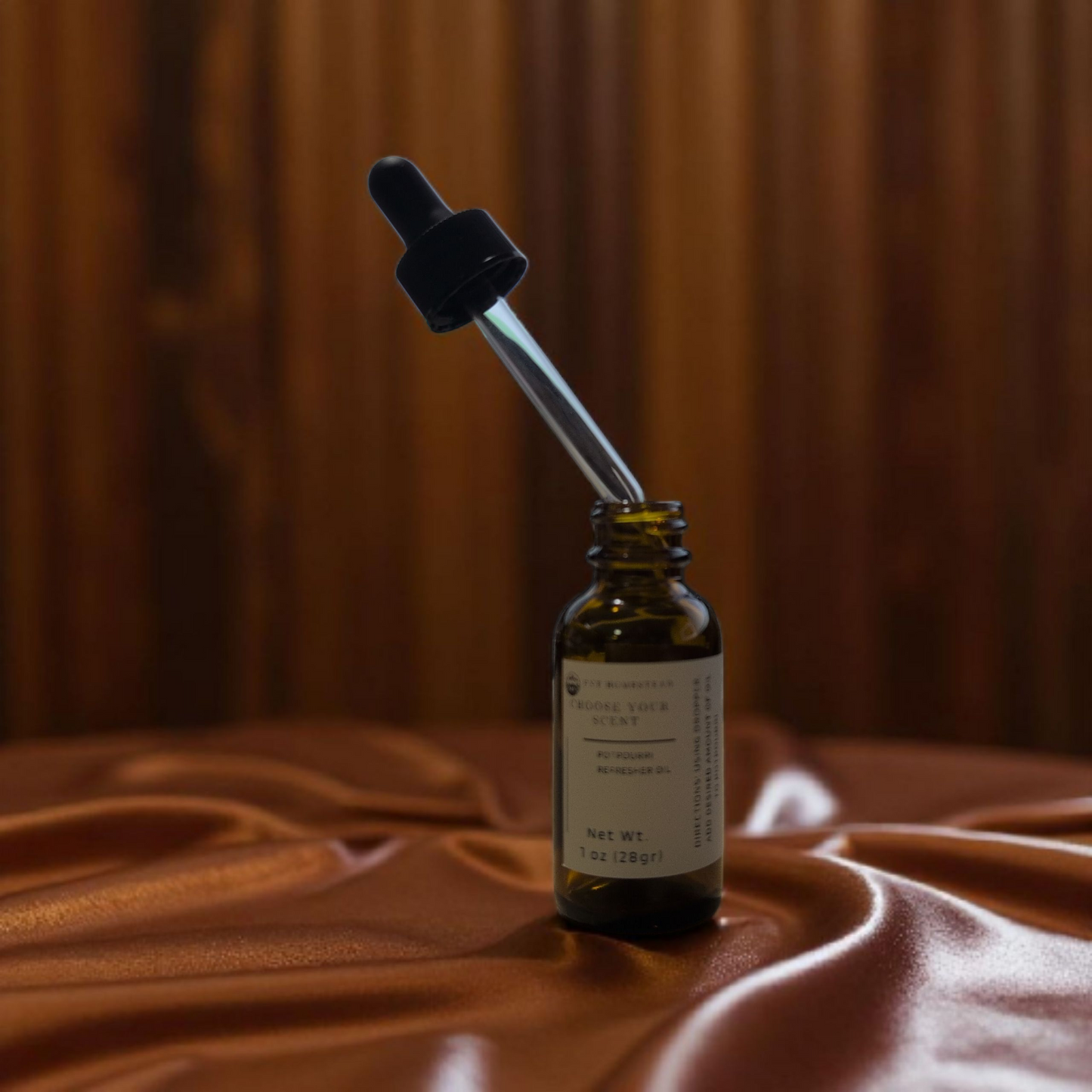 This is an elegant, close-up product photograph with a warm and luxurious feel.
An open, 1 oz. amber glass bottle of "Potpourri Refresher Oil" is the central focus. The bottle rests on shimmering, coppery-brown satin fabric that is gathered in soft folds.
The black dropper has been lifted from the bottle and is held at an angle above it, with the clear glass pipette appearing to hold some of the oil. The background is a continuation of the same rich, silky fabric, draped to create a soft, pleated curtain.