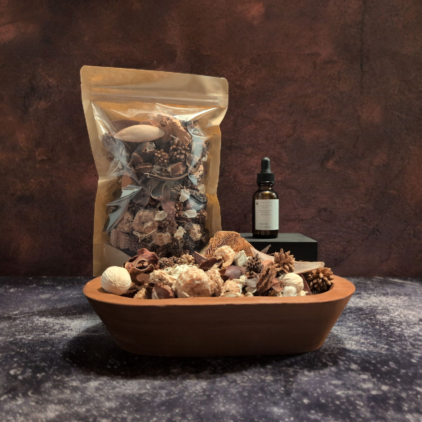 A still life arrangement of a potpourri set sits on a dark, textured surface against a deep brown, moody backdrop. In the foreground, a low terracotta bowl is filled with dried botanicals. Behind it is a stand-up pouch of potpourri and a bottle of potpourri refresher oil.