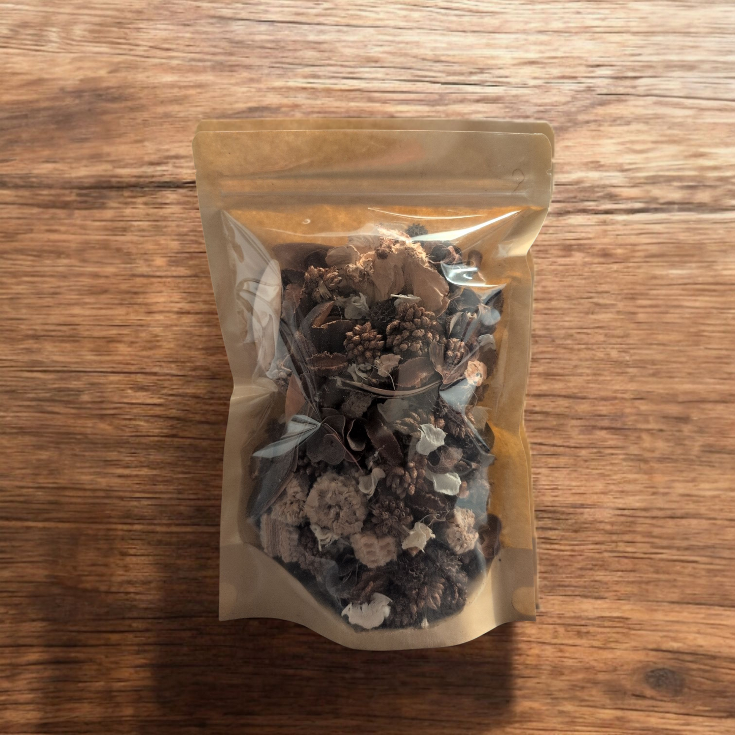 A stand-up pouch with a clear front and brown paper back, filled with potpourri, standing on a wooden surface.