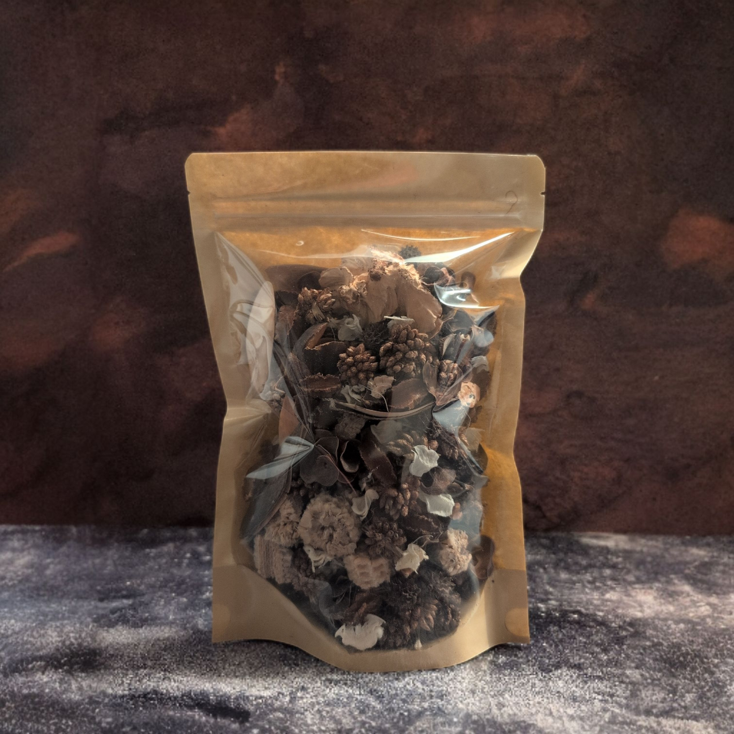 A stand-up resealable pouch with a brown kraft paper back and a clear front window is filled with potpourri. The pouch stands on a dark, slate-like surface against a mottled, dark brown background.