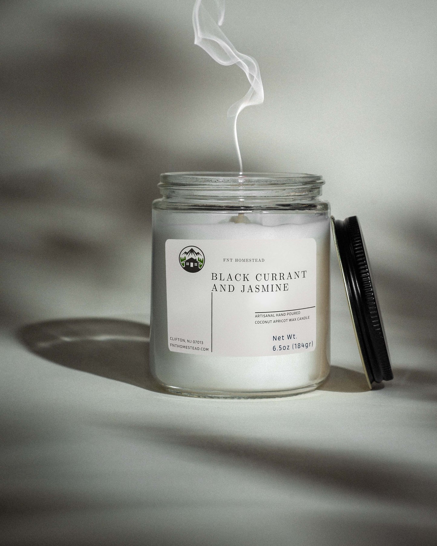 A clear glass jar candle with a black lid, labeled 'Black Currant and Jasmine'.  A wisp of smoke rises from the extinguished wick, suggesting a recently burned candle.