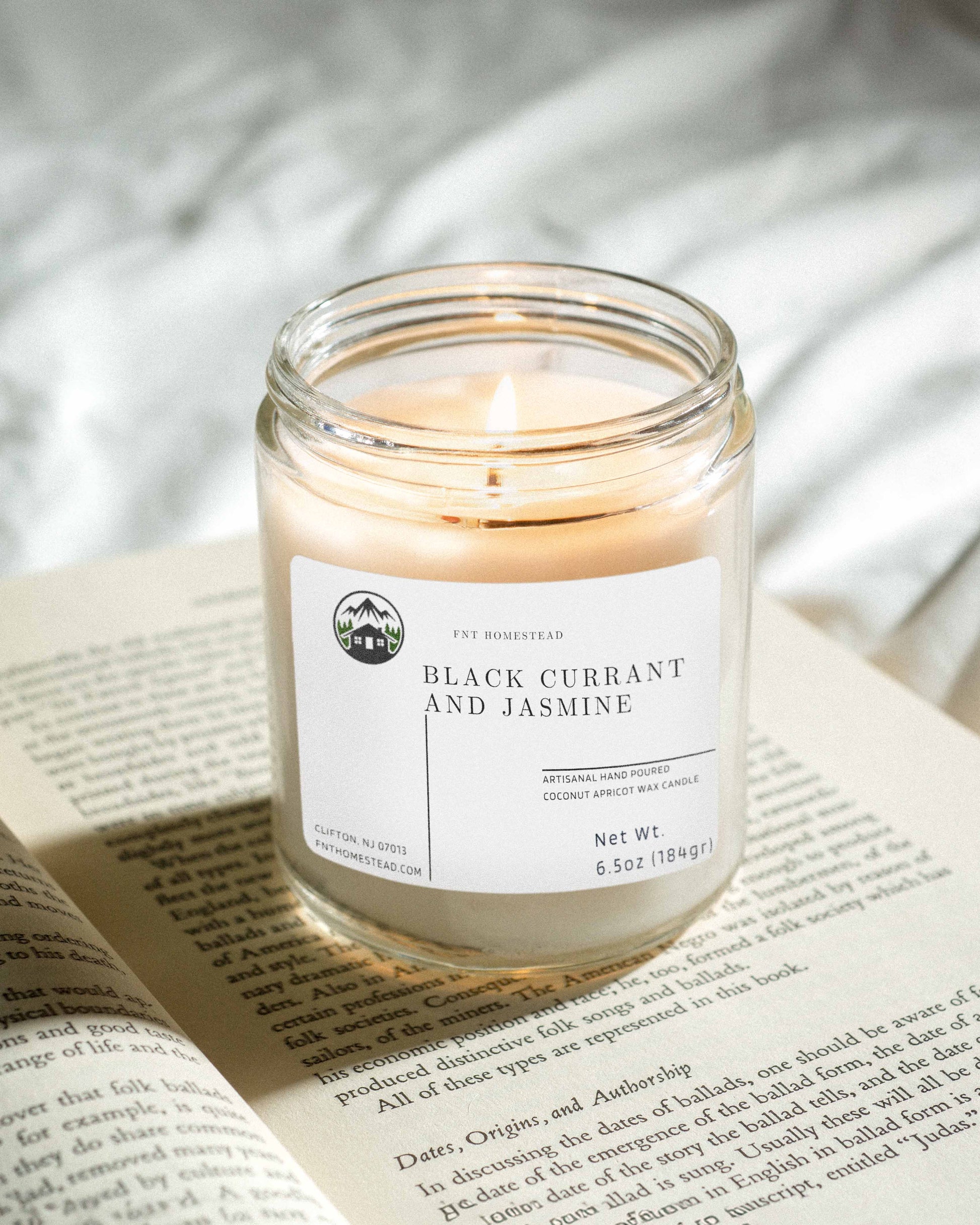 A 'Black Currant and Jasmine' candle with a soft, glowing flame illuminates an open book. The candle sits in a clear glass jar, creating a cozy and inviting atmosphere.