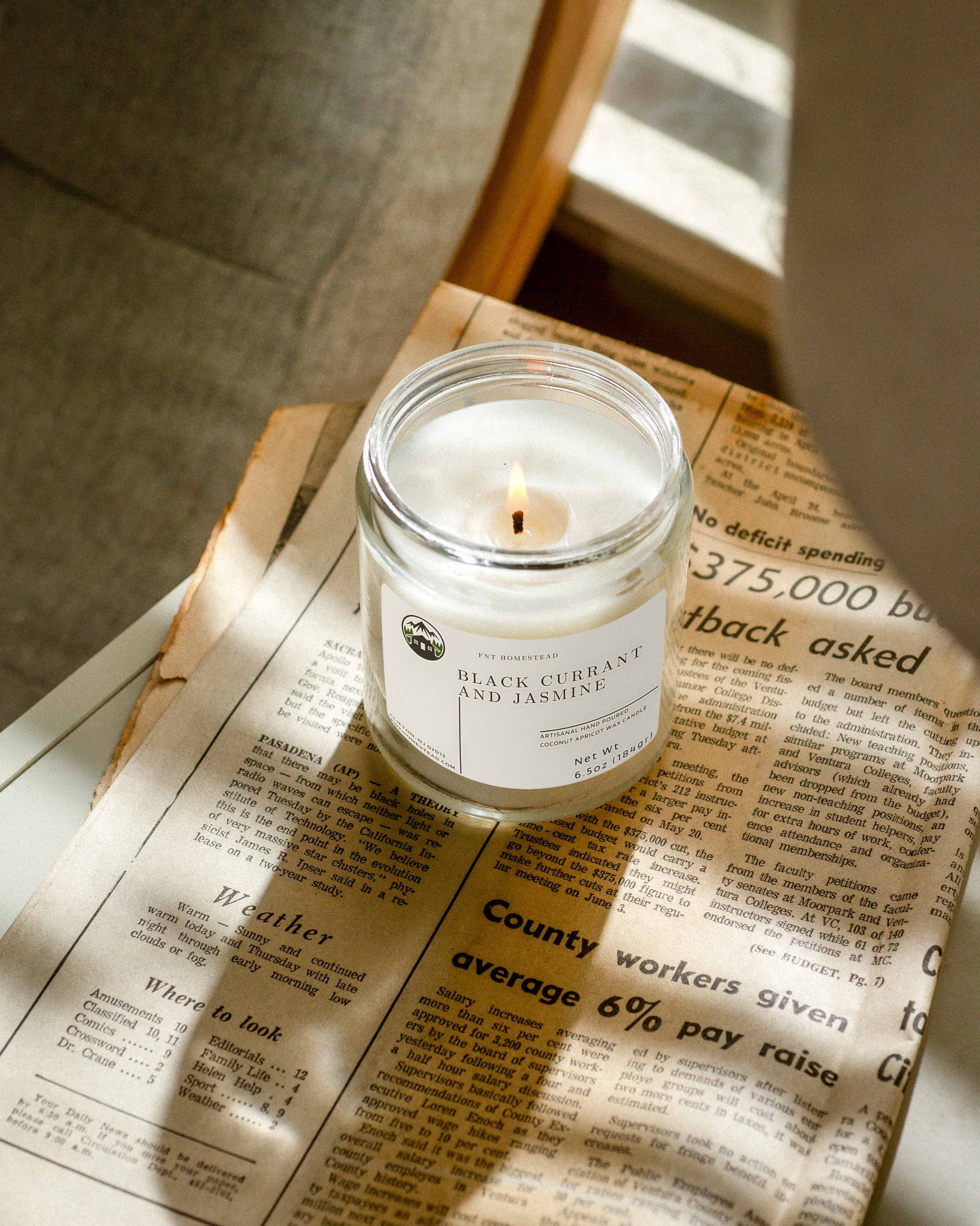 Lit 'Black Currant and Jasmine' candle in a clear glass jar resting on a newspaper, casting warm light on the surrounding text.