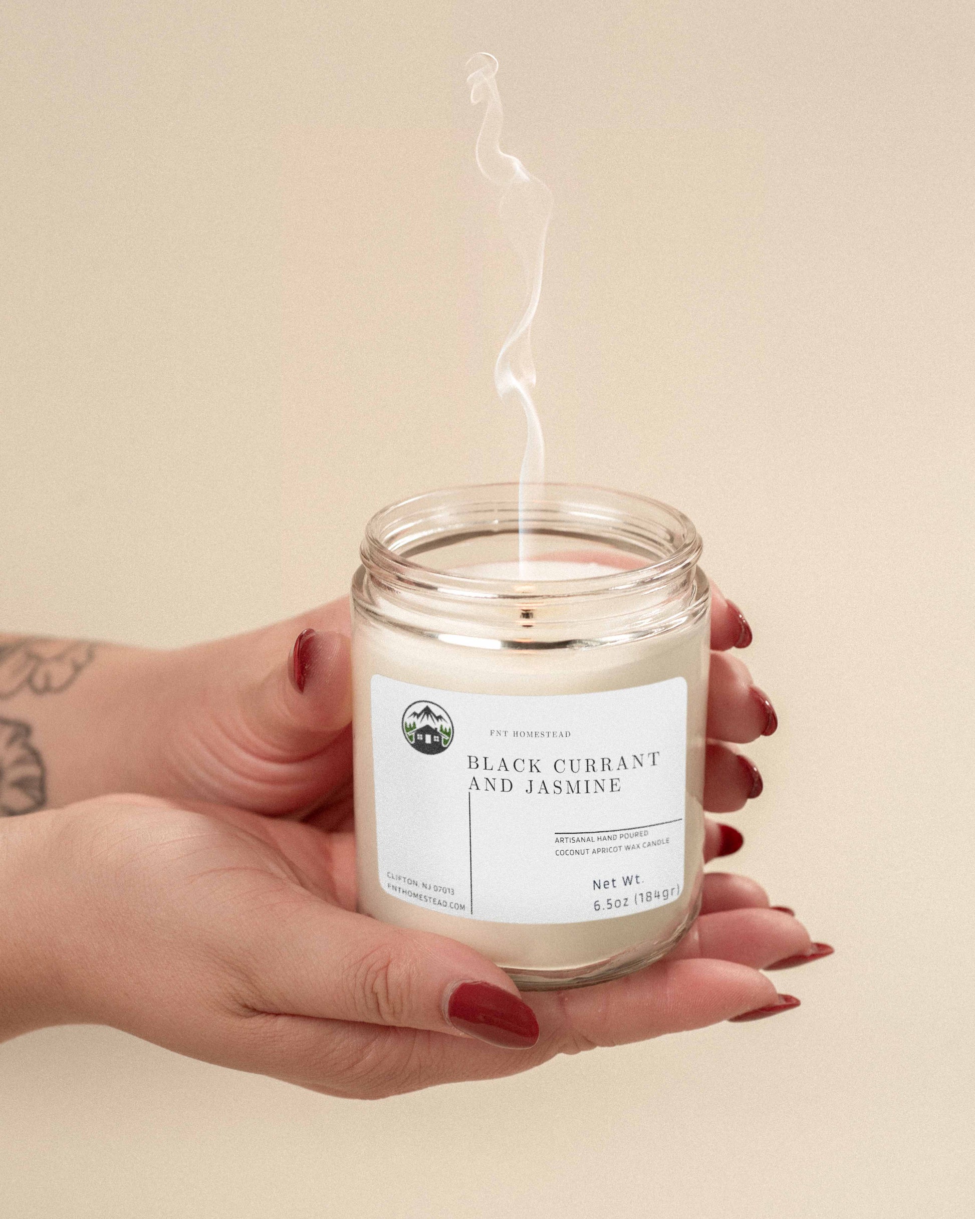 Two hands gently hold a 'Black Currant and Jasmine' candle. The clear glass jar reveals a white wax candle with a single wick, and a delicate wisp of smoke suggests a recently extinguished flame.