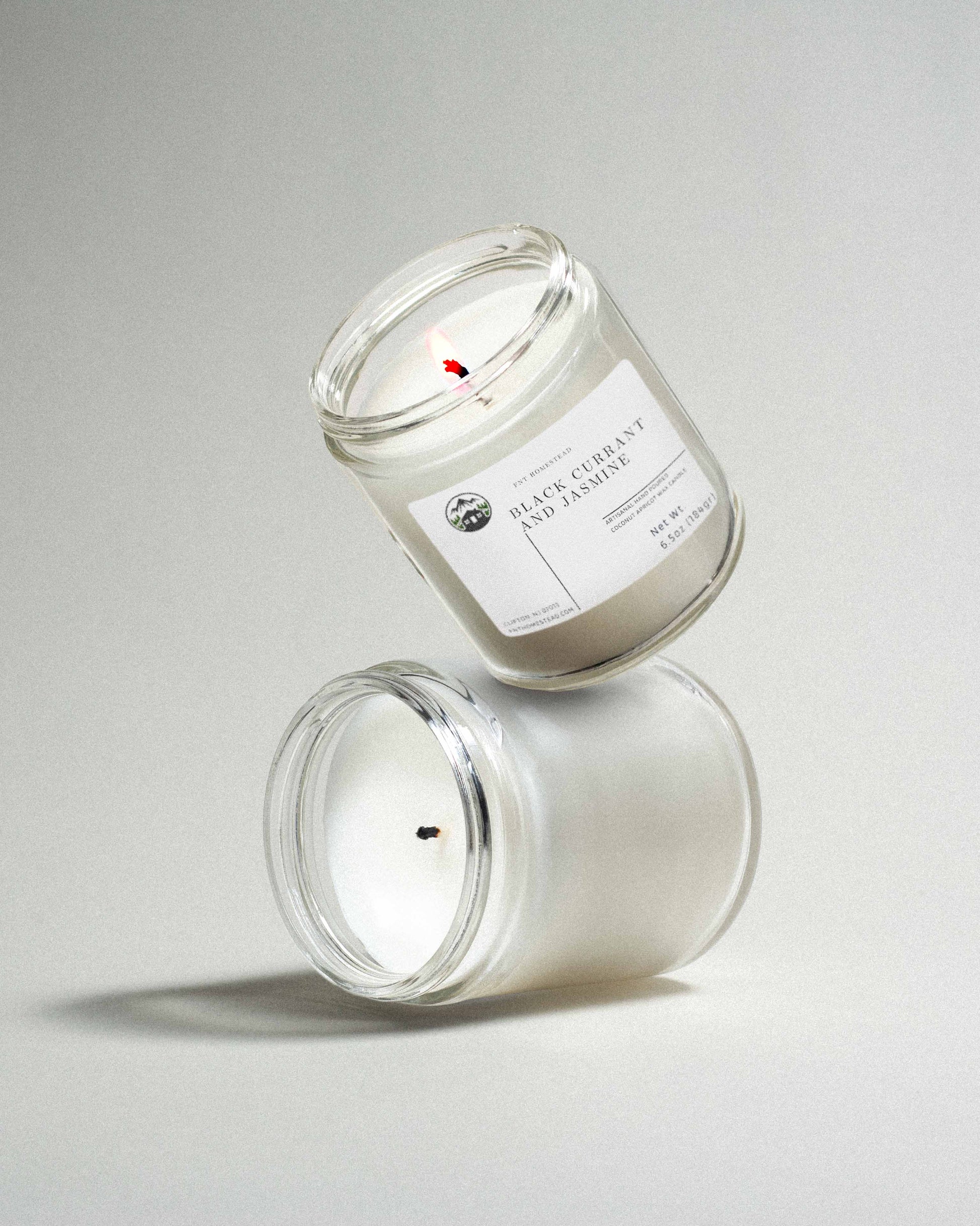 Two clear glass jar candles labeled 'Black Currant and Jasmine', one upright and one tilted standing on top of the upright in a playful manner, showcasing the clean fragrance and minimalist design.