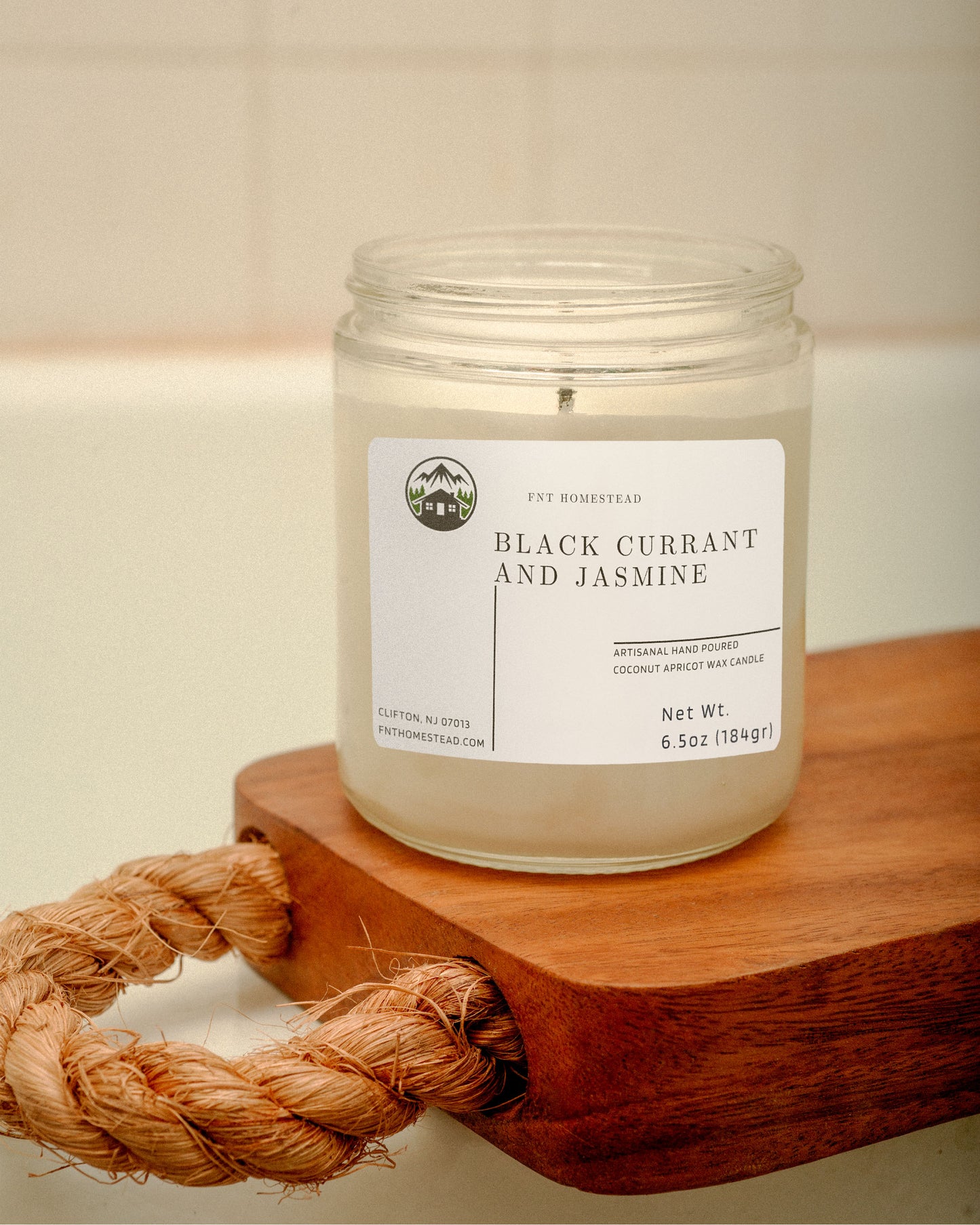 'Black Currant and Jasmine' candle in a clear glass jar with a black lid. The candle is placed on a wooden surface with a coiled rope beside it. Its minimalist design complements any decor.