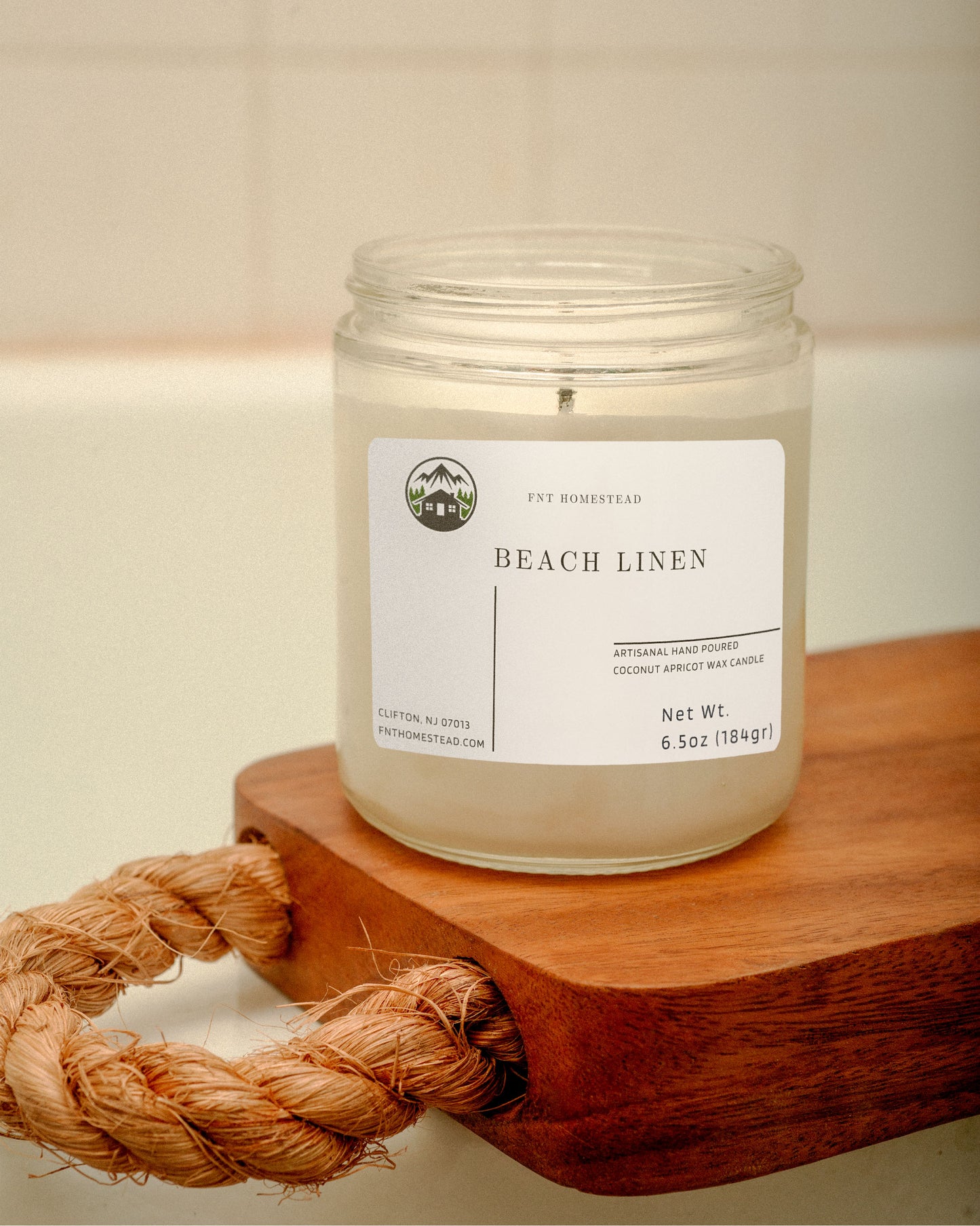 'Beach Linen' candle in a clear glass jar with a black lid. The candle is placed on a wooden surface with a coiled rope beside it. Its minimalist design complements any decor.