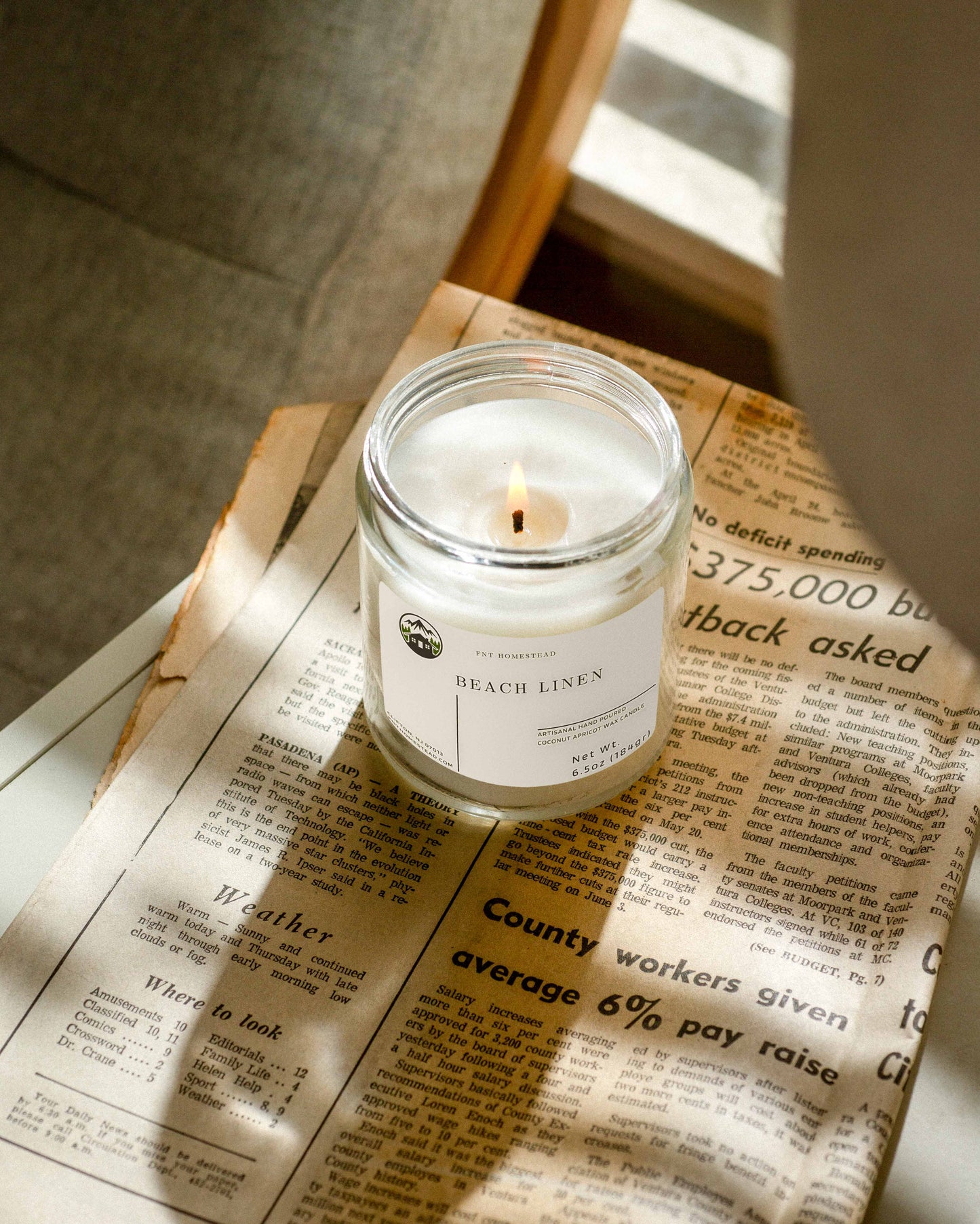 Lit 'Beach Linen' candle in a clear glass jar resting on a newspaper, casting warm light on the surrounding text.