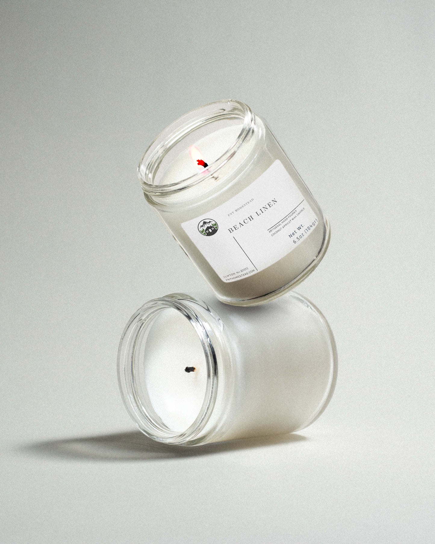 Two clear glass jar candles labeled 'Beach Linen', one upright and one tilted standing on top of the upright in a playful manner, showcasing the clean fragrance and minimalist design.