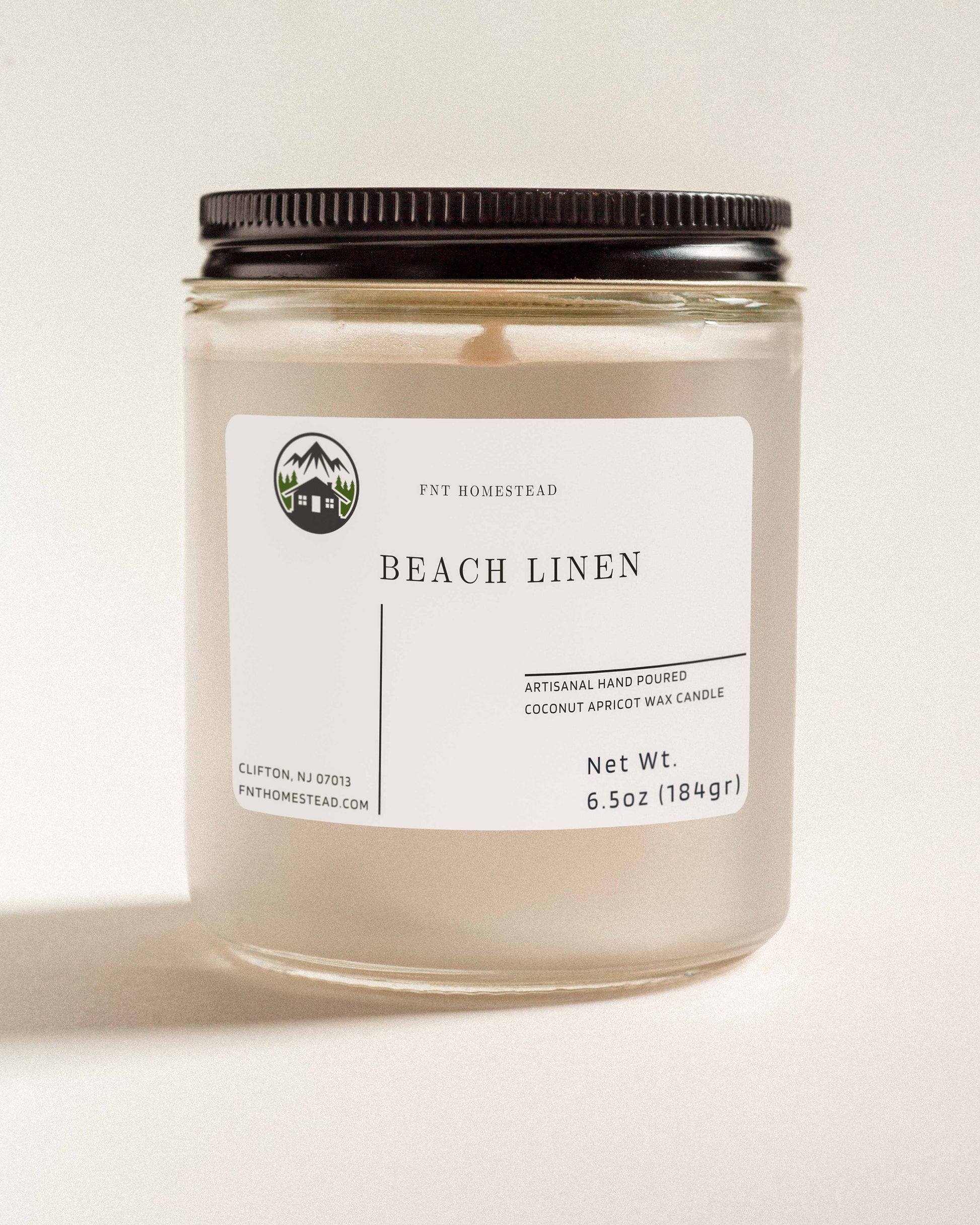 'Beach Linen' scented candle in a clear glass jar with a black lid. This handcrafted candle features a smooth, white wax surface and is made with coconut apricot wax.