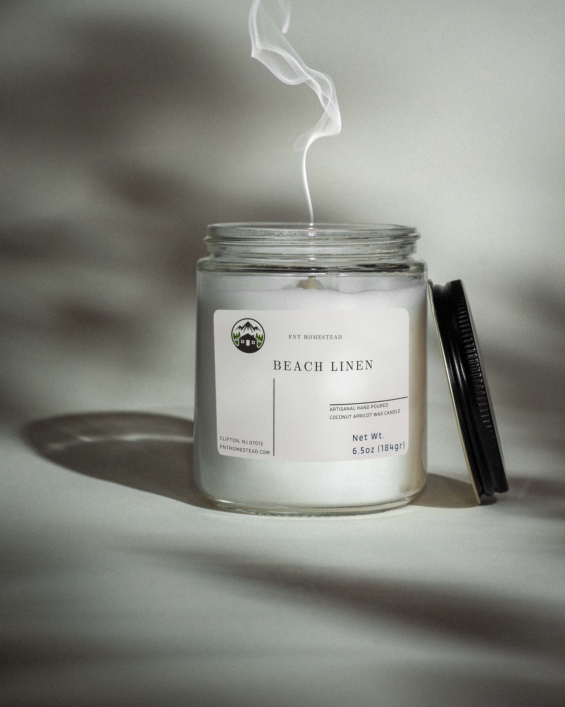 A clear glass jar candle with a black lid, labeled 'Beach Linen'.  A wisp of smoke rises from the extinguished wick, suggesting a recently burned candle.