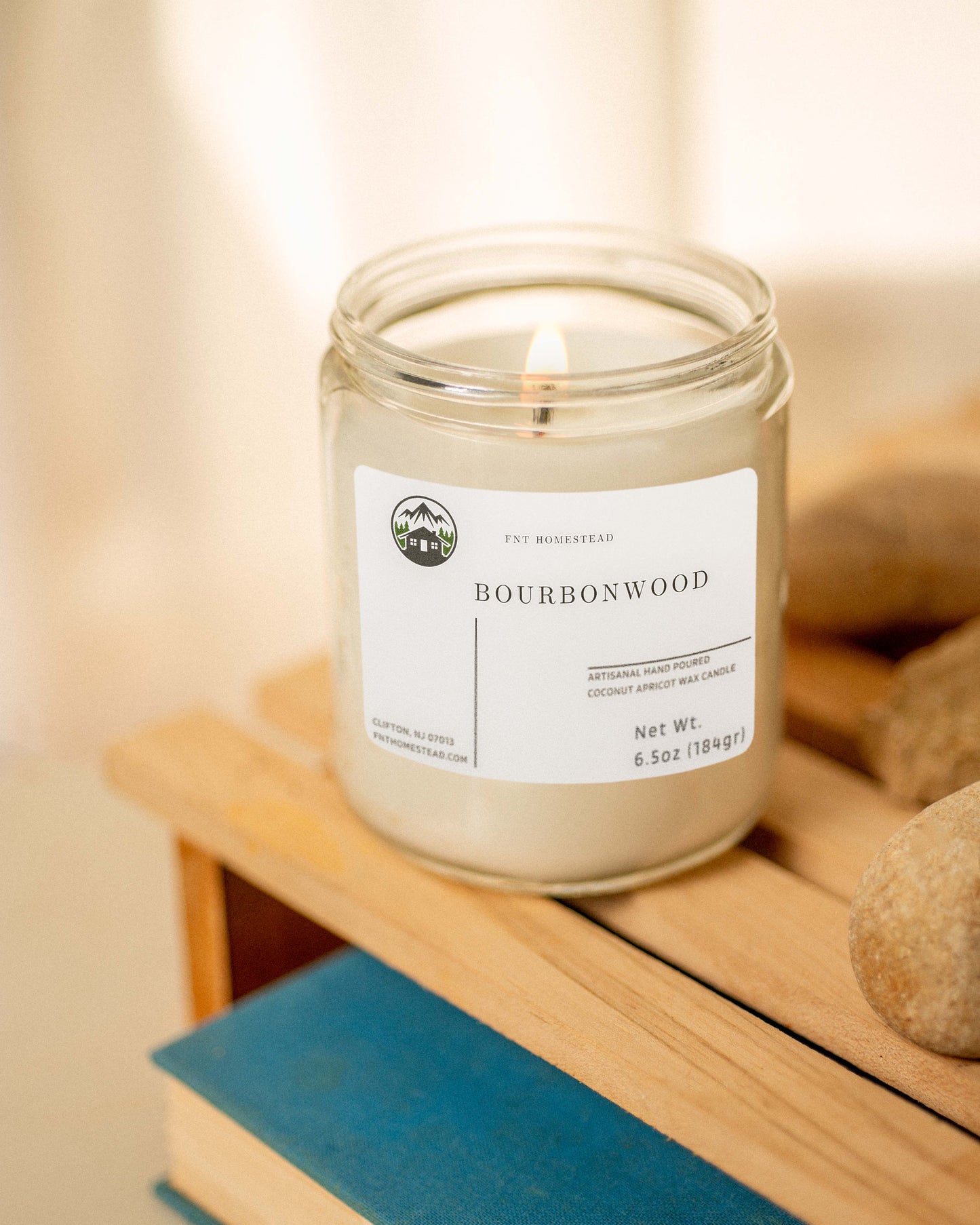 The 'Bourbonwood' candle creates a relaxing atmosphere with its soft glow in this clear glass jar, placed on a wooden surface with a book and stones.