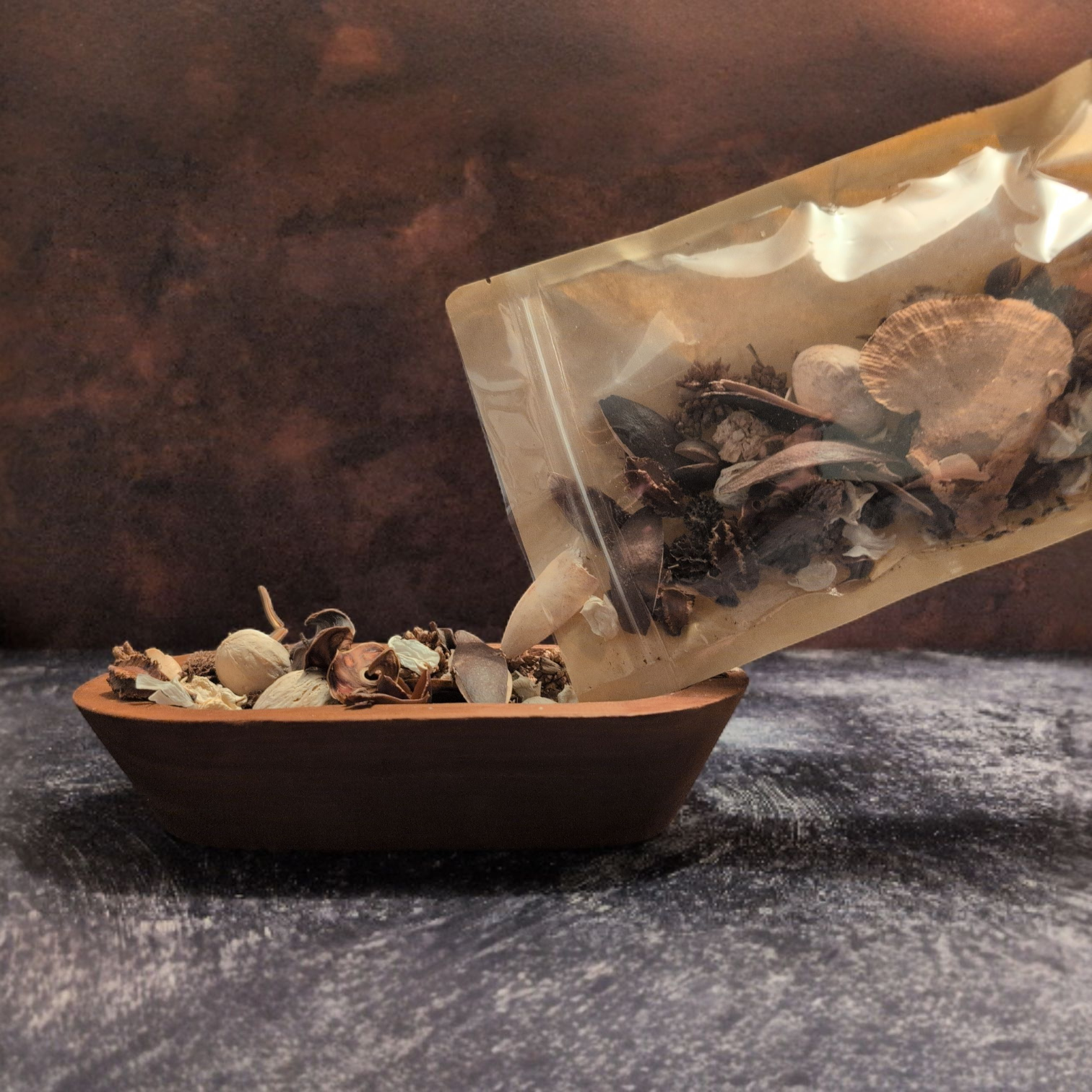 A hand pours potpourri from a clear and brown paper pouch into a shallow, dark brown bowl. The bowl and pouch are set against a dark, textured background of brown and gray.