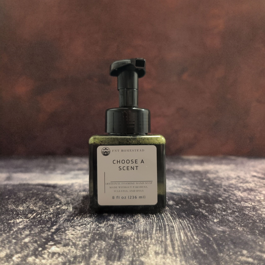 A square, dark green bottle of FNT Homestead foaming hand soap is centered in the photo against a dark, moody background. The soap inside the bottle has a golden shimmer, and the bottle sits on a dark, textured surface. The white label on the front reads, "Choose a Scent."