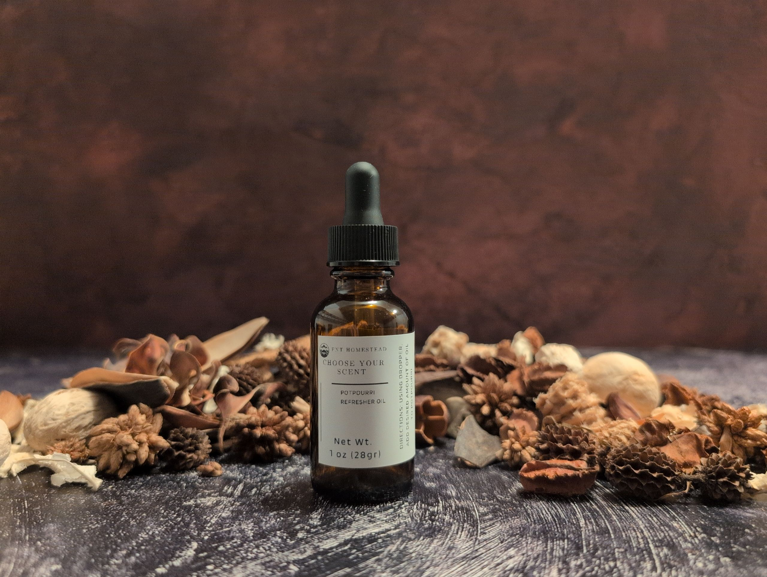 A 1 oz. amber glass bottle with a black dropper cap is centered in the frame, nestled in a bed of dried, earthy-toned potpourri. The white label on the bottle reads "CHOOSE YOUR SCENT" and "POTPOURRI REFRESHER OIL." The scene is set on a dark, textured surface against a deep brown backdrop, creating a warm and rustic mood.

